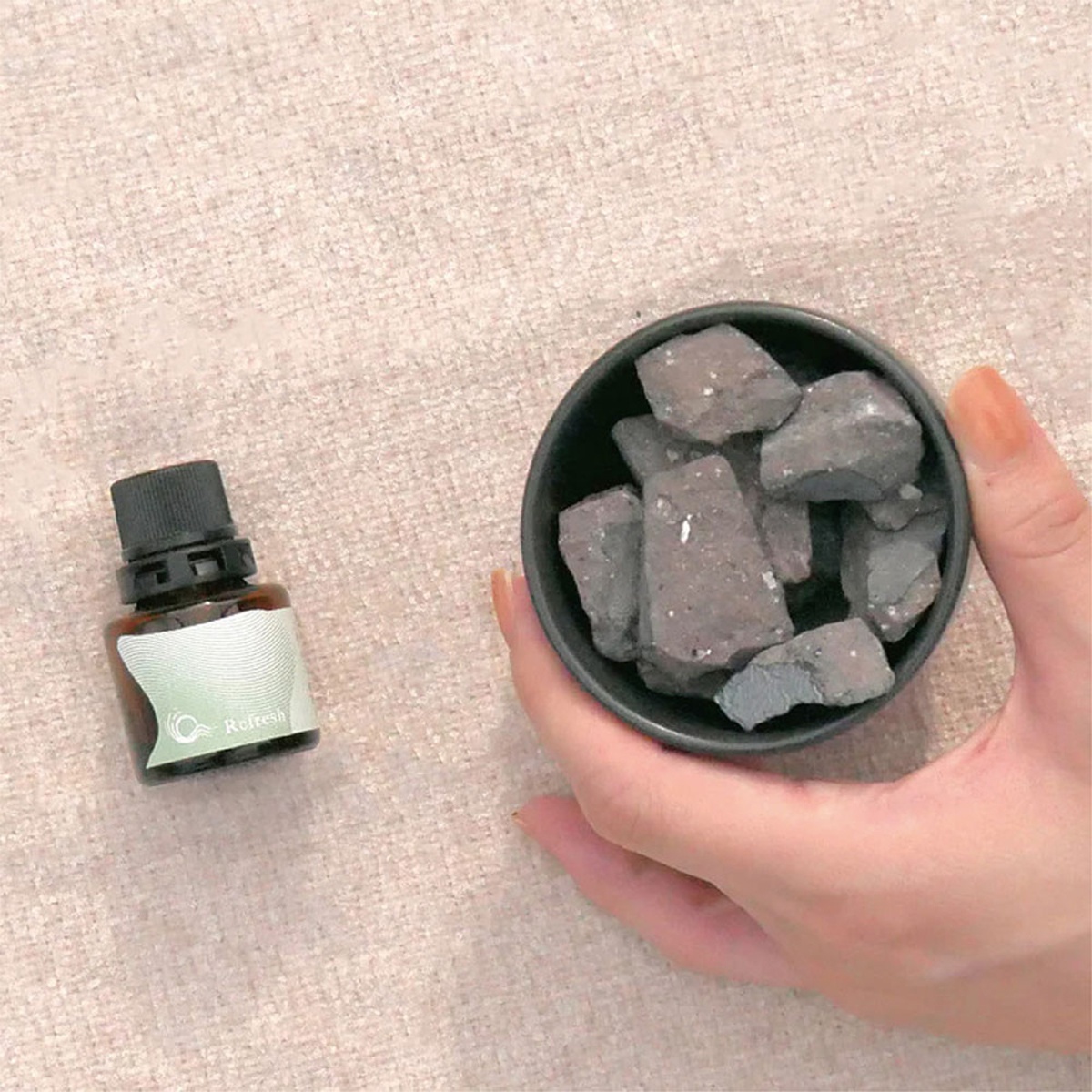 一瓶標有 Refresh 的香薰精油旁，一隻手拿著一個裝有灰色擴香石的小碗。