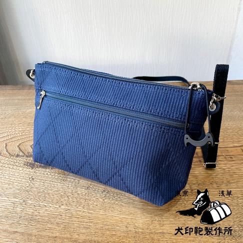 深藍色刺子織紋理斜背包，正面有兩條拉鍊，側邊附有黑色織帶和一個黑色吊飾。