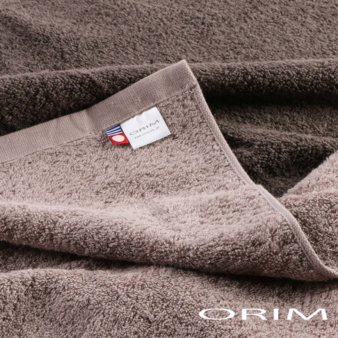 BULKY PRO今治沙龍浴巾，採用柔軟的純棉材質，呈現深淺兩種大地色調，毛圈蓬鬆，質感厚實。浴巾一角縫有品牌標籤，標籤上印有ORIM字樣及網址，並帶有紅藍色條紋裝飾。