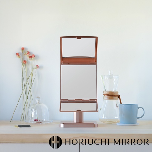 玫瑰金色桌上型三折化妝鏡，鏡面可360度旋轉，附有底座，擺放在木質檯面上，背景為白色牆面，旁有乾燥花束、玻璃罩盅、咖啡壺和藍色馬克杯。