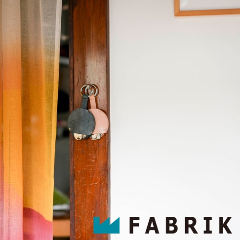 FABRIK 品牌標誌旁有兩個皮革製的圓形收納鑰匙圈，一個是深灰色，另一個是粉紅色，懸掛在木門上，左側是色彩鮮豔的窗簾。