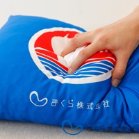 藍色布面小抱枕，印有白色愛心圖案與日文「まくら株式会社」字樣，抱枕中央有紅色與藍白色條紋的圓形標誌，手正按壓抱枕，展現其柔軟材質。