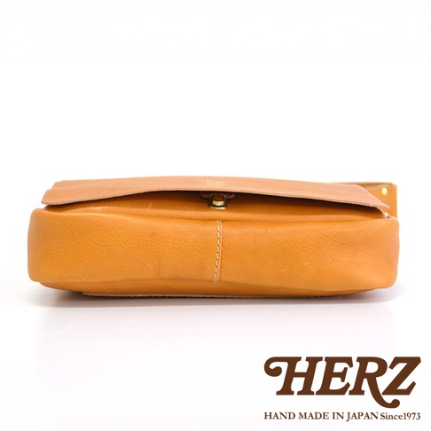 HERZ 品牌標誌，下方為 HAND MADE IN JAPAN Since1973 字樣，圖片主體為一個棕褐色皮革側背包的側面特寫，包身呈現圓潤飽滿的造型。