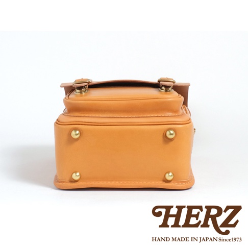 職人手作皮革小學生書包，呈現經典的棕褐色皮革，設計簡約，底部設有四個黃銅保護釘，頂部有金屬釦環和皮革帶，整體風格復古耐用。