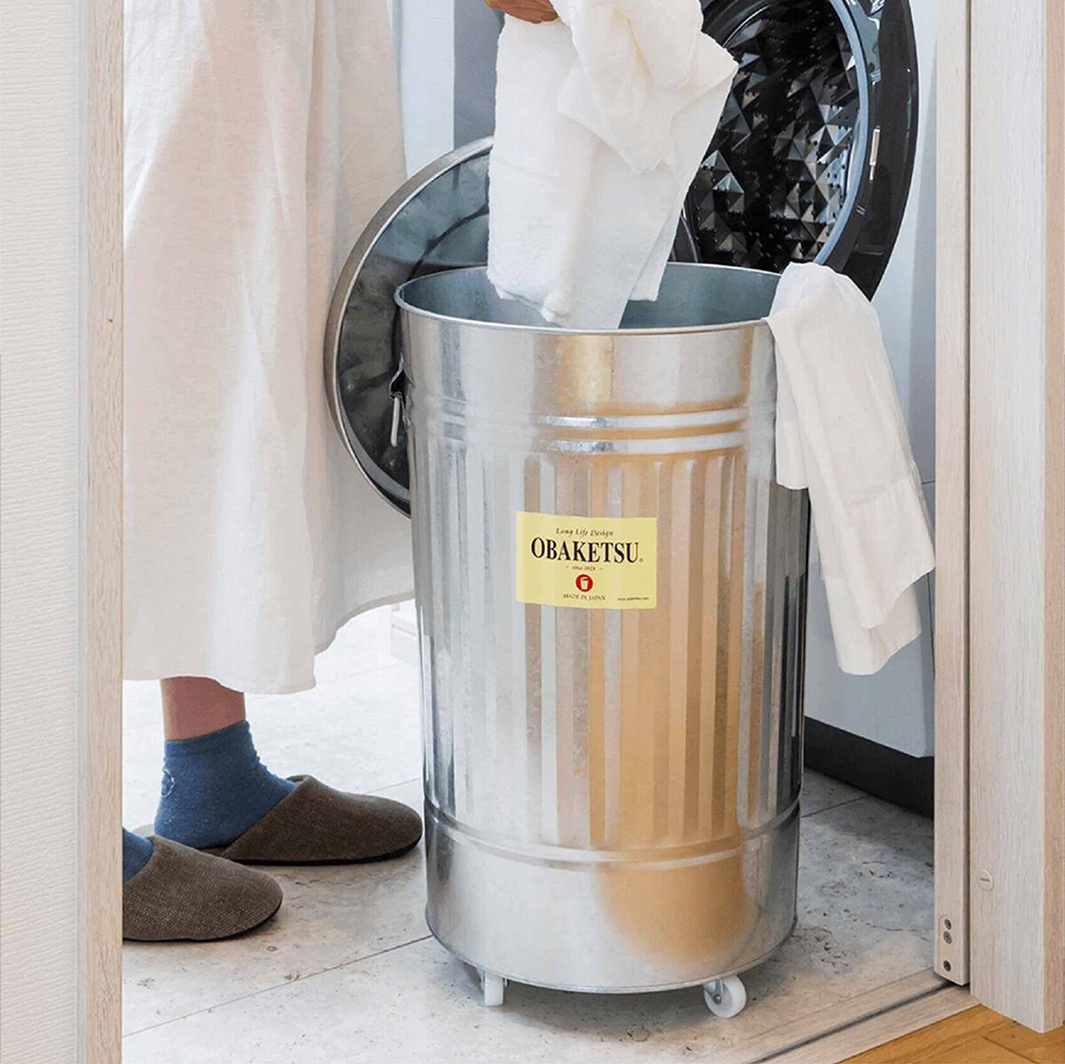 一個戴著藍色襪子和棕色拖鞋的人，正將一件白色衣物放入一個帶有 OBAKETSU 標籤的鍍鋅洗衣籃中。