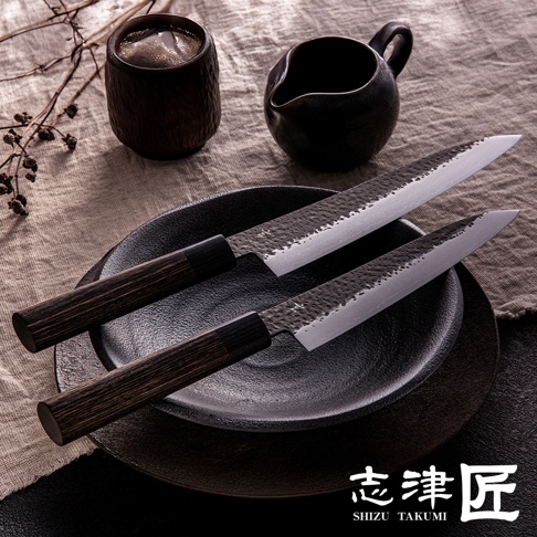 兩把「玄 / Chef's knife 主廚刀21cm」，刀刃呈現鍛造紋理，握柄為深木色，置於黑色圓形盤中，背景為亞麻布，旁有一黑色茶杯與小茶壺。