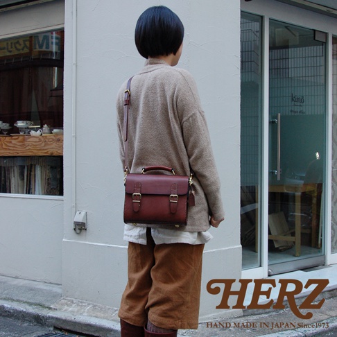 一款深棕色皮革製成的極簡風格迷你單肩包，具有兩個金屬扣環的翻蓋設計，配有頂部提把和可拆卸肩帶，可斜挎或肩背。