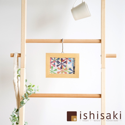 木質牆面收納架，懸掛著一個淺木色相框，框內是色彩繽紛的花朵圖案。架子上方掛著一個白色馬克杯，左側懸掛著一個米白色帆布袋，架子旁有綠色植物點綴。