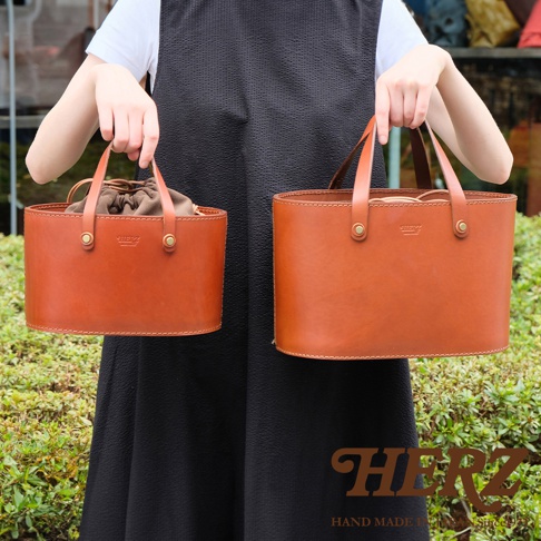 兩款職人手作皮革束口手提包，皆為溫暖的棕褐色，採用光滑皮革材質，搭配同色系束口內袋與皮質提把，設計簡約大方。