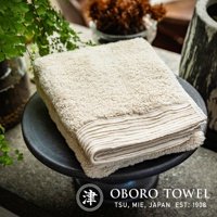 一條米白色、觸感蓬鬆的毛巾，帶有羅紋邊飾，放置於黑色圓形底座上。