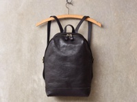 深棕色皮革後背包，簡約褶皺設計，配有雙拉鍊主袋，皮革材質，掛於木質衣架上，背景為淺色紋理牆面。