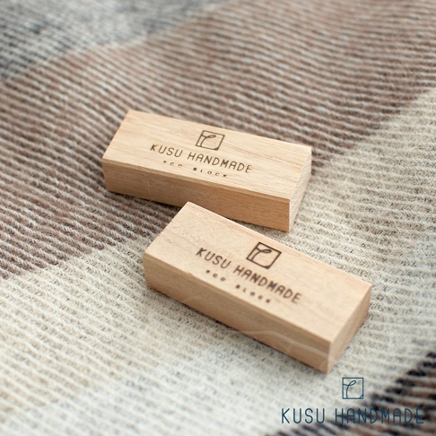 兩塊淺色木質長方條，表面印有黑色「KUSU HANDMADE ECO BLOCK」文字及品牌標誌，放置於紋理織物上。