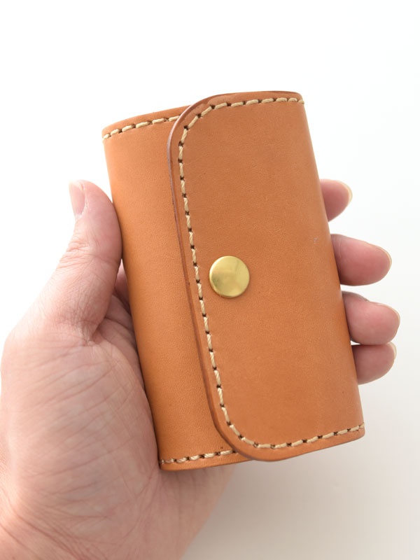 手持一個棕褐色皮革製的經典鑰匙包，包身以手工縫線裝飾，中央有一個金色按扣。