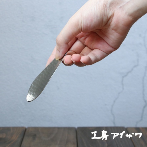 手持一把銀色不鏽鋼茶勺，勺身呈寬扁葉片狀，表面有錘紋，勺頭圓潤。