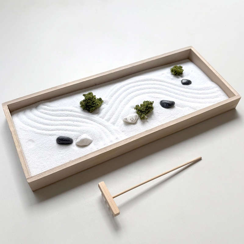 一個長方形木製盒裝的日式枯山水模型，內部鋪滿細緻白砂，砂面由隨附的小木耙畫出流暢的波浪紋理。盒中裝飾著幾個深色鵝卵石、白色小石塊以及幾簇綠色植栽，呈現出靜謐的微型庭園景觀。
