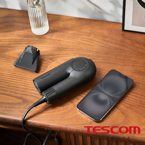 一個魅影黑的TESCOM摺疊式吹風機，附有集風嘴，旁放著一支手機，擺放在木紋桌面上。