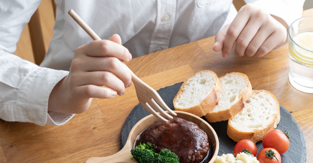 淺色楓木餐叉，三齒設計，用於叉取漢堡排，搭配麵包、花椰菜、小番茄與檸檬水。