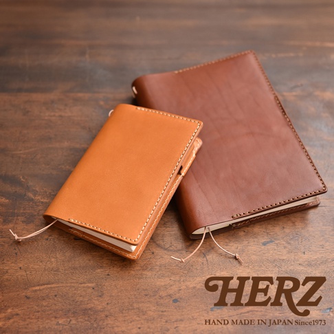 近景為一個淺棕色皮革筆記本套，後面有一個深棕色皮革筆記本套，皆有縫線細節，置於木紋背景上。