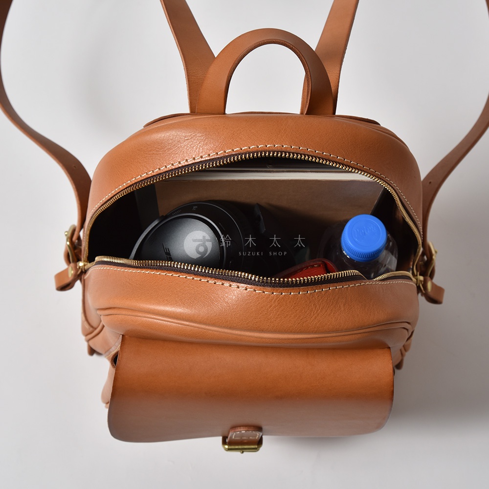 棕色皮革迷你後背包，立體前袋，拉鍊開口可見內部裝有耳機、書本和水瓶。