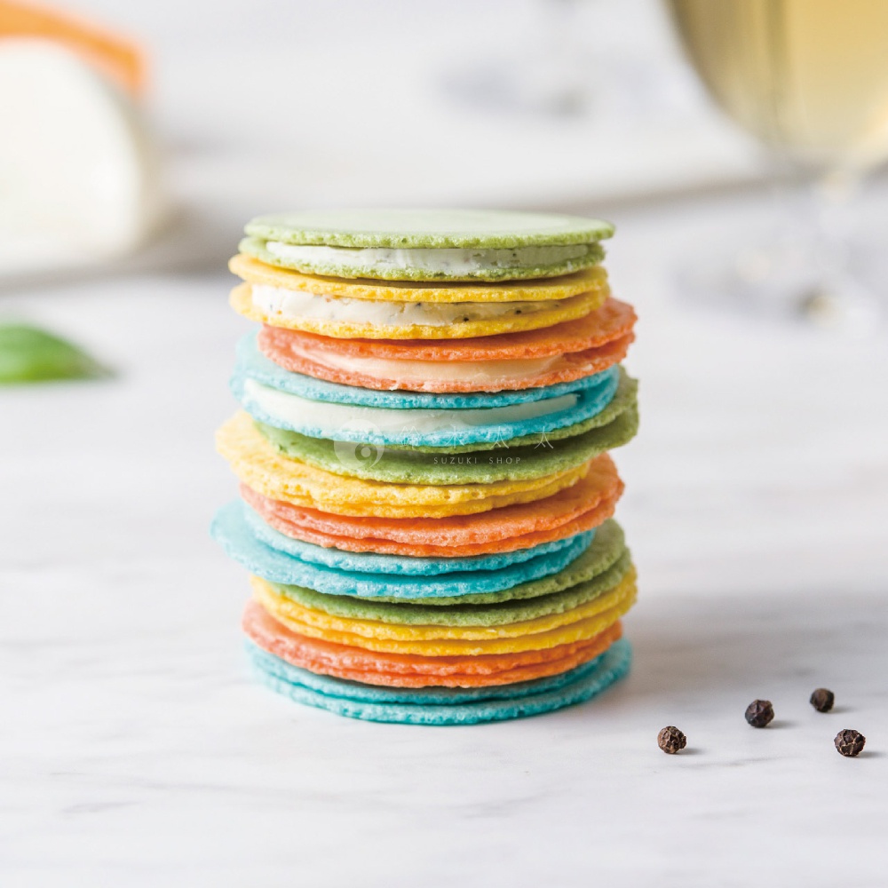 在淺色的雲石紋桌面上，堆疊著一座色彩繽紛的馬卡龍造型起司蝦仙貝塔，呈現出青綠、鮮黃、橘紅與天藍色交替的層次感，仙貝之間夾著細緻的乳酪內餡，畫面右下方散落著幾顆黑色胡椒粒，整體風格明亮簡約，展示出精緻的點心質感。