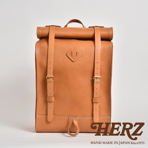 一個 tan 色全皮革後背包，採用捲口設計，配有兩個可調節的肩帶和黃銅扣環。包包正面中央有一個菱形縫線裝飾，底部有一個皮革拉環，整體風格簡潔。