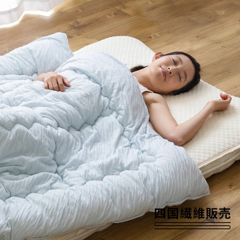 一位女性蓋著淺藍色舒芙蕾透氣四季被（中棉款），被子表面有明顯的褶皺紋理，鋪在米白色菱格紋床墊上，背景是木地板。