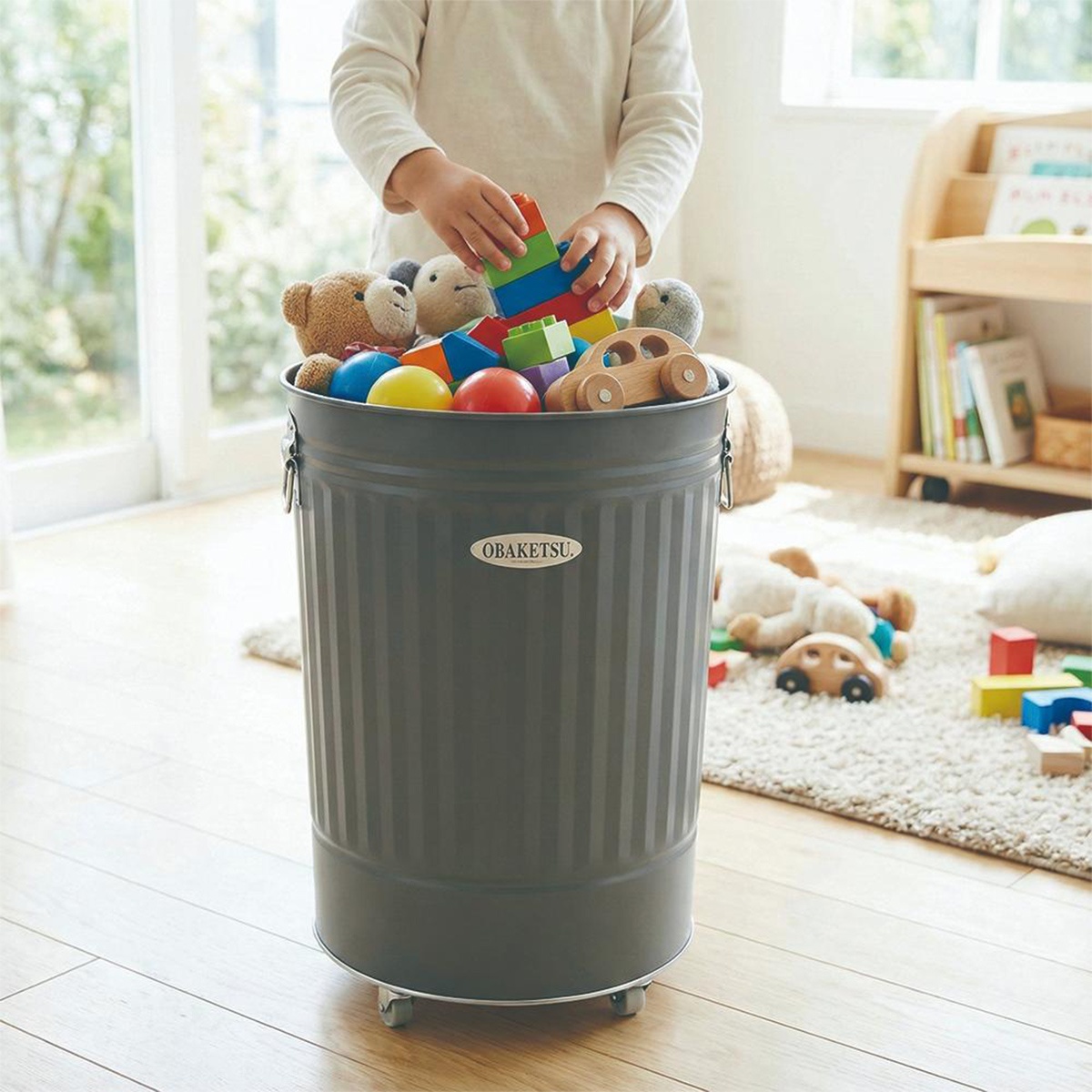 OBAKETSU 品牌的灰色附輪垃圾桶，裝滿了各種玩具，包含積木、絨毛玩具和木製小汽車，一個小孩正將積木放入桶中。