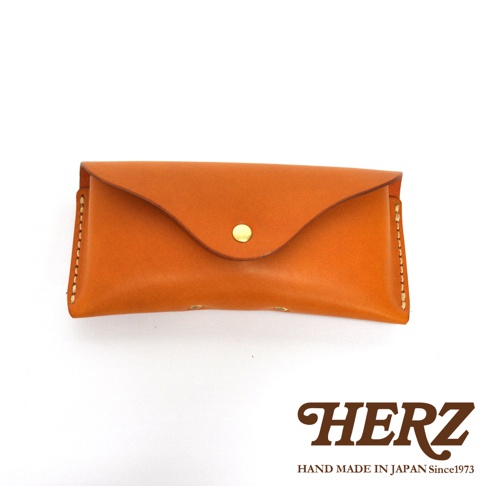 手工製作的橘棕色皮革眼鏡套，配有金色摁扣，邊緣縫線清晰可見，下方印有HERZ品牌標誌。
