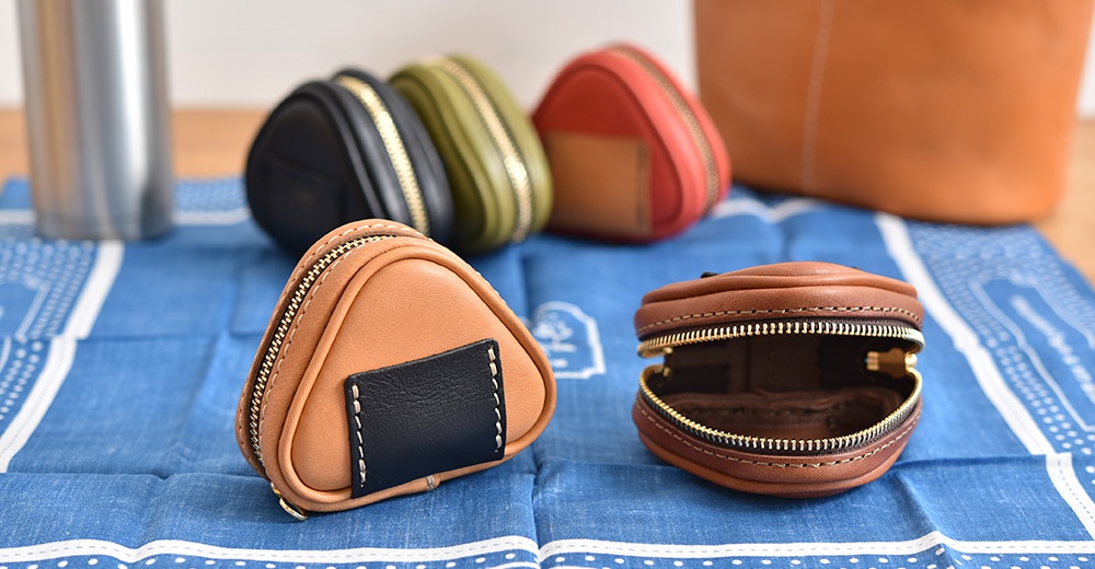 多個職人手作皮革三角飯糰造型小物收納包，顏色包含棕色、黑色、綠色、紅色。材質為皮革，配有金色拉鍊。其中一個棕色收納包打開展示內部。