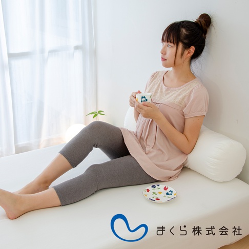 一位女性坐在床上，雙腿夾著一個白色圓柱形枕頭。她身穿粉色上衣和灰色七分褲，手中拿著一個印有花朵圖案的杯子，側身望向窗外。