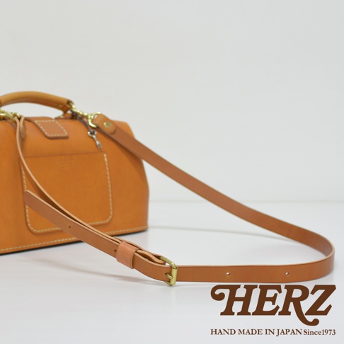 職人手作皮革長型迷你醫生包，採用天然皮革材質，呈現溫潤的焦糖棕色。包款設有手提握把及可調節長度的肩背帶，搭配復古金屬釦件，整體風格簡約而精緻。