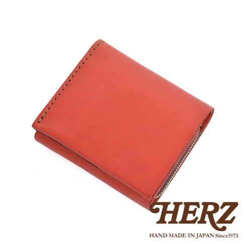 一個日本HERZ手工製作的紅色皮革雙折短夾，採用柔軟的原創牛皮革製成，表面呈現細膩的質地與鮮豔的橘紅色調。皮夾側面有粗獷精緻的縫線細節，並配有L型拉鍊零錢袋，整體設計簡約俐落，右下角印有HERZ品牌標誌及手工製造資訊。