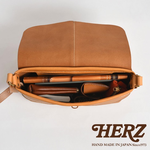 HERZ 職人手作皮革細扣帶馬鞍包，從上方視角展示打開的包內皮革物品，包含筆、皮夾及零錢包，下方是品牌 LOGO。