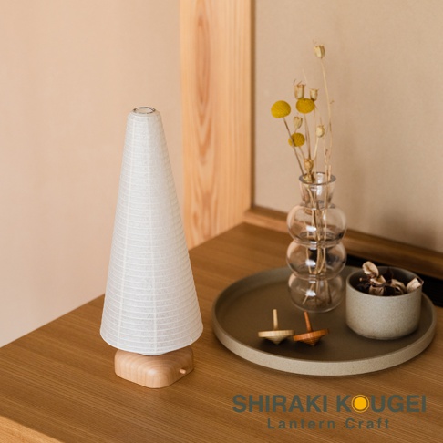 錐形白色紙質提燈，帶有細緻的網格紋理，底部為淺木色底座。提燈旁是裝有乾燥花草的透明玻璃花瓶，及一個灰色圓形托盤，上面有兩個木製陀螺。