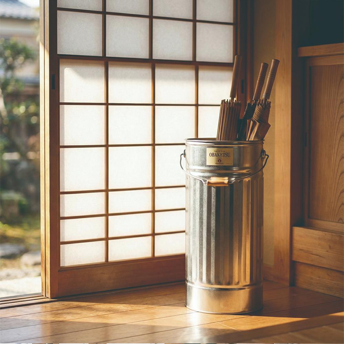 日式空間中，一個 OBAKETSU 品牌的手工金屬傘桶，裝著幾把木柄的傘，靠在紙拉窗旁，陽光灑落在木質地板上。