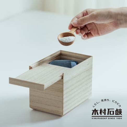 手持木製湯匙，舀取白色洗衣粉，倒入淺色木盒中。木盒蓋子部分開啟，盒內可見藍色包裝的產品。