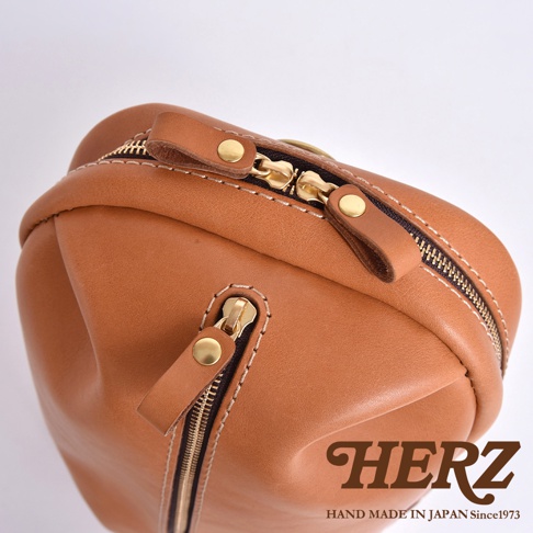 日本 HERZ 品牌手工製作的棕色皮革立體運動斜背包，特寫鏡頭展示其高品質的皮革材質、精緻的縫線以及閃亮的金色拉鍊與五金配件，背包上印有 HERZ 品牌標誌及「HAND MADE IN JAPAN Since1973」的文字。