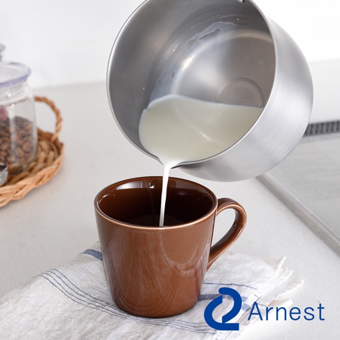 不鏽鋼萬用鍋傾倒牛奶至深棕色馬克杯中，杯子放置於條紋布上，背景有裝有咖啡豆的玻璃罐。