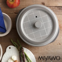 灰色陶土湯鍋，鍋蓋上飾有白色網格線，中央設有圓形把手及排氣孔，鍋身側面為黑色，置於木質桌面，旁有紅蘋果、藍色餐巾、白色餐盤及蔬菜。