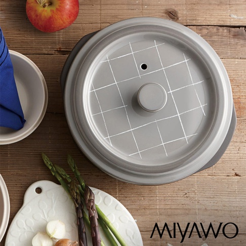 灰色陶土湯鍋，鍋蓋上飾有白色網格線，中央設有圓形把手及排氣孔，鍋身側面為黑色，置於木質桌面，旁有紅蘋果、藍色餐巾、白色餐盤及蔬菜。