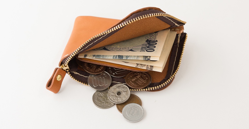 一個淺棕色皮革製成的迷你拉鍊錢包，頂部開口處放置了一張一千元日圓紙鈔，錢包下方散落著幾枚不同面值的日本硬幣，呈現出精緻簡約的職人手作質感。