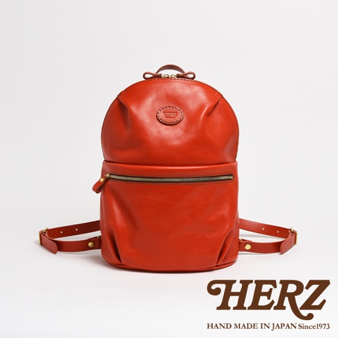 橘紅色皮革製成的大開口圓角後背包，正面有一個拉鍊口袋，頂部有提把和品牌標誌。
