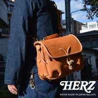 咖啡色皮革製成的職人手作鹿角扣繩郵差包，包身有自然壓紋，搭配淺棕色皮革滾邊與提把，金色金屬釦環與鹿角造型拉繩，側面有鉚釘裝飾。