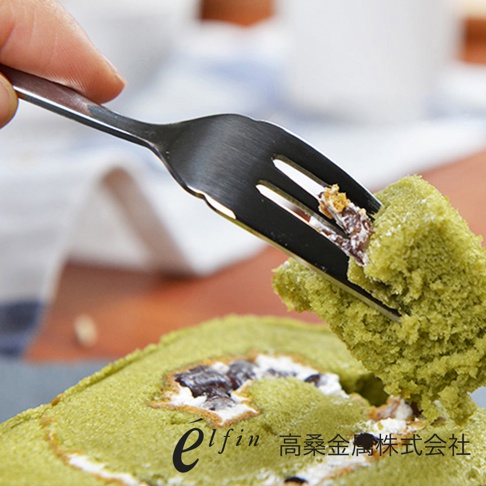 一只手持著日製錘目紋不鏽鋼水果甜點叉，叉尖挑起一塊綠色蛋糕。叉子為銀色，材質為不鏽鋼，表面有細緻的錘目紋理。背景模糊，顯示出蛋糕卷和一些餐具。
