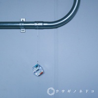 透明水晶立方體吊飾，底部有橙色和藍色光澤，懸掛於金屬管下方，背景為淺藍色牆面。