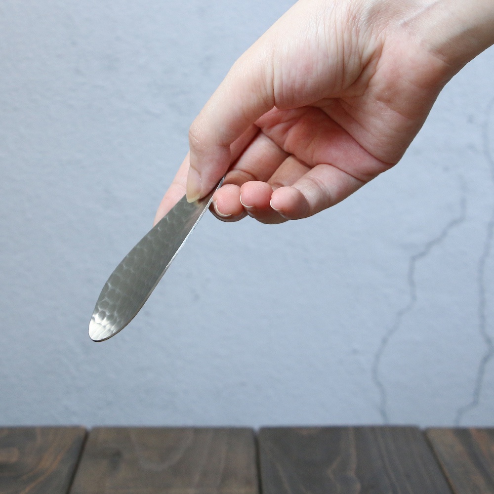 一只手拿著一把銀色不鏽鋼茶勺，勺面有錘紋紋理，勺柄細長。