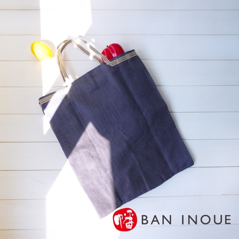 深藍色蚊帳棉織隨身購物袋，兩側有條紋織帶裝飾，袋口露出兩個紅色的水果模型，左側有一個黃色水果模型。