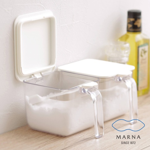 兩個透明塑膠調味料盒，每個約600ml容量，裝有白色粉末。盒蓋為白色，其中一個盒蓋打開。塑膠提把透明，方便拿取。背景為白色牆壁與木質桌面，右側有黃色瓶裝液體。