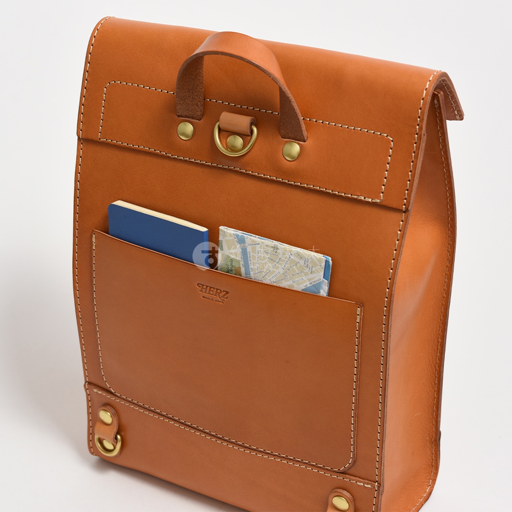 棕褐色皮革製的紙袋風後背包，正面有一個大口袋，內有藍色筆記本和地圖。包包採用精細縫線，配有頂部提把和金屬扣環，整體呈現簡約質感。