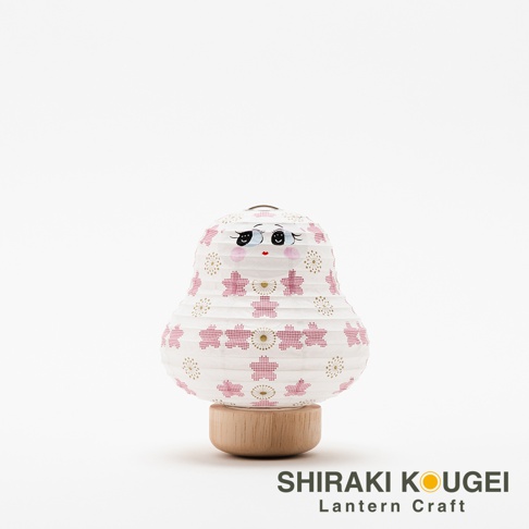 白色紙質達摩不倒翁迷你提燈，燈身印有粉紅色櫻花圖案與金色放射狀紋飾，臉部有可愛的黑色眼睛、紅色嘴唇和粉紅色臉頰，底部為木質座。